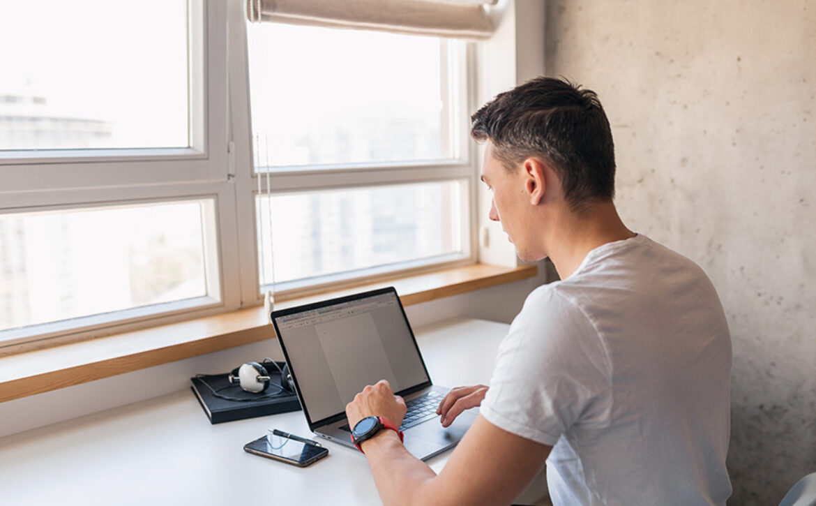 young handsome man in casual outfit sitting at table working on laptop, freelancer at home, view from back