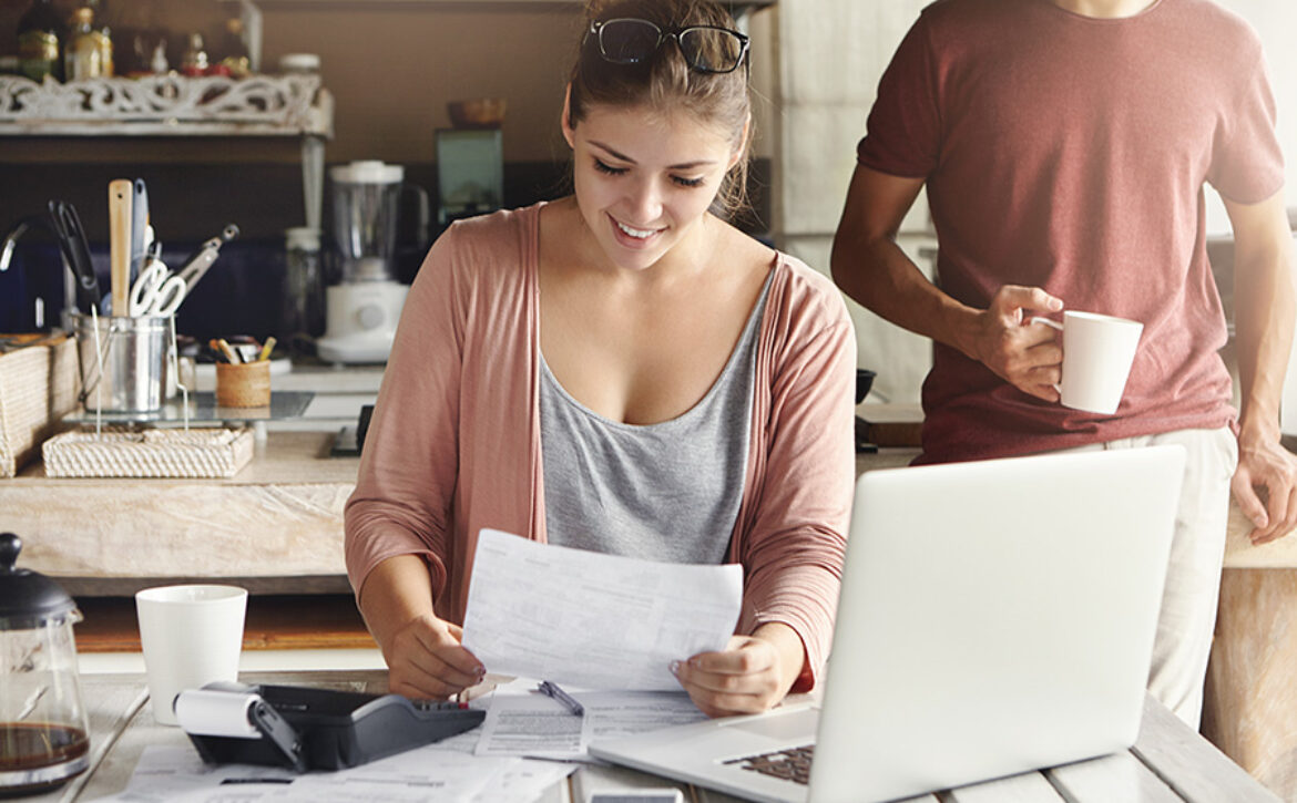 Young pretty woman wearing glasses on her head smiling happily while reading document saying that bank approved their mortgage application, her husband standing on background and drinking coffee